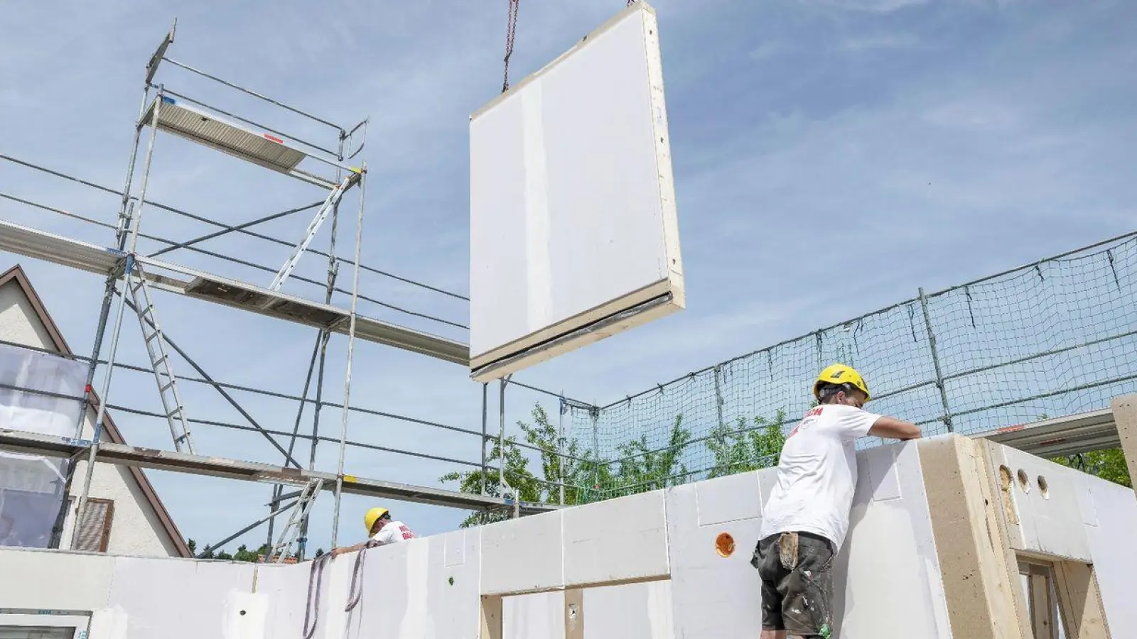 Ein Bauarbeiter steht auf einer Leiter und arbeitet im Rohbau eines Fertighauses, während gerade ein weiteres Wandelement von einem Kran heruntergelassen wird. (Foto: Daniel Maurer/dpa)