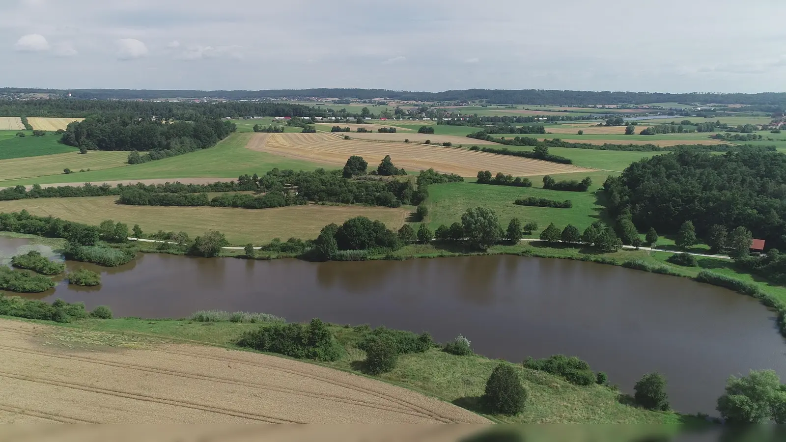 Der Stausee bei Dorfgütingen mit dem Festival-Gelände: Der Zugangsbereich wird nach gegenwärtiger Planung unweit der Fischerhütte (rechts) aufgebaut. Davor ist ein öffentlich zugänglicher Biergarten geplant, an dessen Betrieb örtliche Vereine schon ihr Interesse bekundet haben. Die Grünfläche links von dem Waldstück ist als Campingplatz und Backstage-Bereich vorgesehen. Derweil soll die Bühne bei der Hecke aufgestellt werden, die die beiden Wiesen voneinander abgrenzt. Als Parkplatz ist ein im Hintergrund zu sehendes Areal ins Auge gefasst. (Foto: Stadt Feuchtwangen/Werner Tippmann)