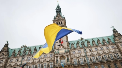 Ukraine-Krieg - Dritter Jahrestag. Ein Mann steht mit ukrainischer Fahne und Trauerflor bei der Gedenkminute „Hamburg steht still“ vor dem Rathaus. (Foto: Christian Charisius/dpa)