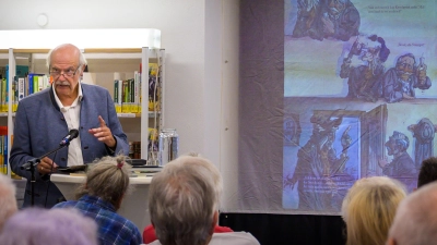Karl-Heinz Röhlin liest in der Gemeindebücherei Neuendettelsau aus seiner fränkischen Fassung des „Brandner Kaspar”. Auf der Leinwand hinter ihm laufen die Zeichnungen von Jan Reiser mit. (Foto: René Chlopotowski)