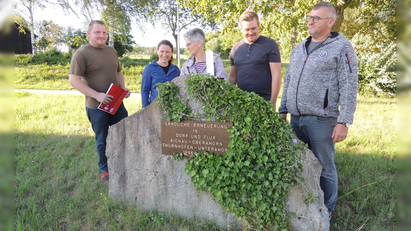 Die Dorfgemeinschaft von Aichau plant den Bau einer Kapelle oberhalb der Böschung im Hintergrund. Das Projekt stellten jetzt Felix Gundacker, Tamara Perske und Edith Geißler sowie Daniel und Martin Gögelein vor, die unser Bild (von links) am Stein zur Erinnerung an die Flurbereinigung zeigt. Auf diesem steht die weiße Kerze, die ihren endgültigen Platz im Innern des Gotteshauses finden soll. (Foto: Peter Zumach)