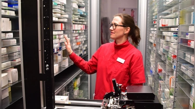 Annette Pauli erklärt, wie Arzneimittel per Zufallsprinzip automatisch auf freie Fächer verteilt werden. Die Packungen werden von dem Schieber zunächst horizontal zu den passenden Etagen gebracht und dann nach oben oder unten vor das per Zufall ausgewählte freie Fach bugsiert. (Foto: Kurt Güner)
