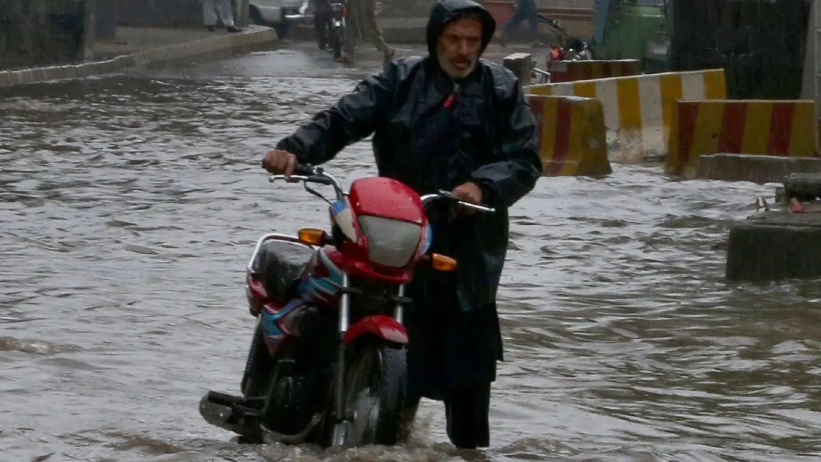 Ein Pakistaner watet nach starken Regenfällen mit seinem Motorrad durch eine überschwemmte Straße in Peschawar. (Foto: Muhammad Sajjad/AP/dpa)