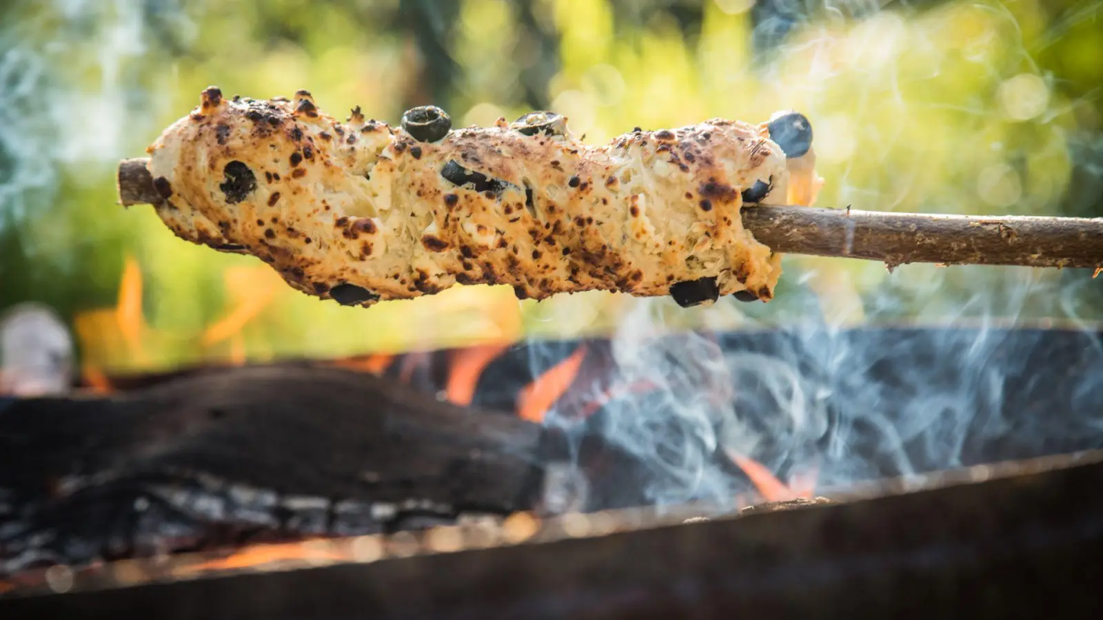 Ein Oliven-Feta-Knoblauch-Hefeteig ist um einen Haselnuss-Stock gewickelt und wird über dem Lagerfeuer gebacken. (Foto: Christin Klose/dpa-tmn)