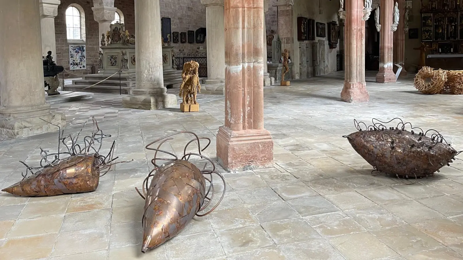 Angelika Summas Wesen aus Kupferblech und Kupferrohr erinnern an riesige Insekten, die im Mortuarium auf dem Rücken gelandet und liegengeblieben sind. Auch die Holzskulpturen im Hintergrund von Andreas Kuhnlein und Harald Kienle schaffen neue Spannungsfelder im Münster Heilsbronn. (Foto: Lara Hausleitner)