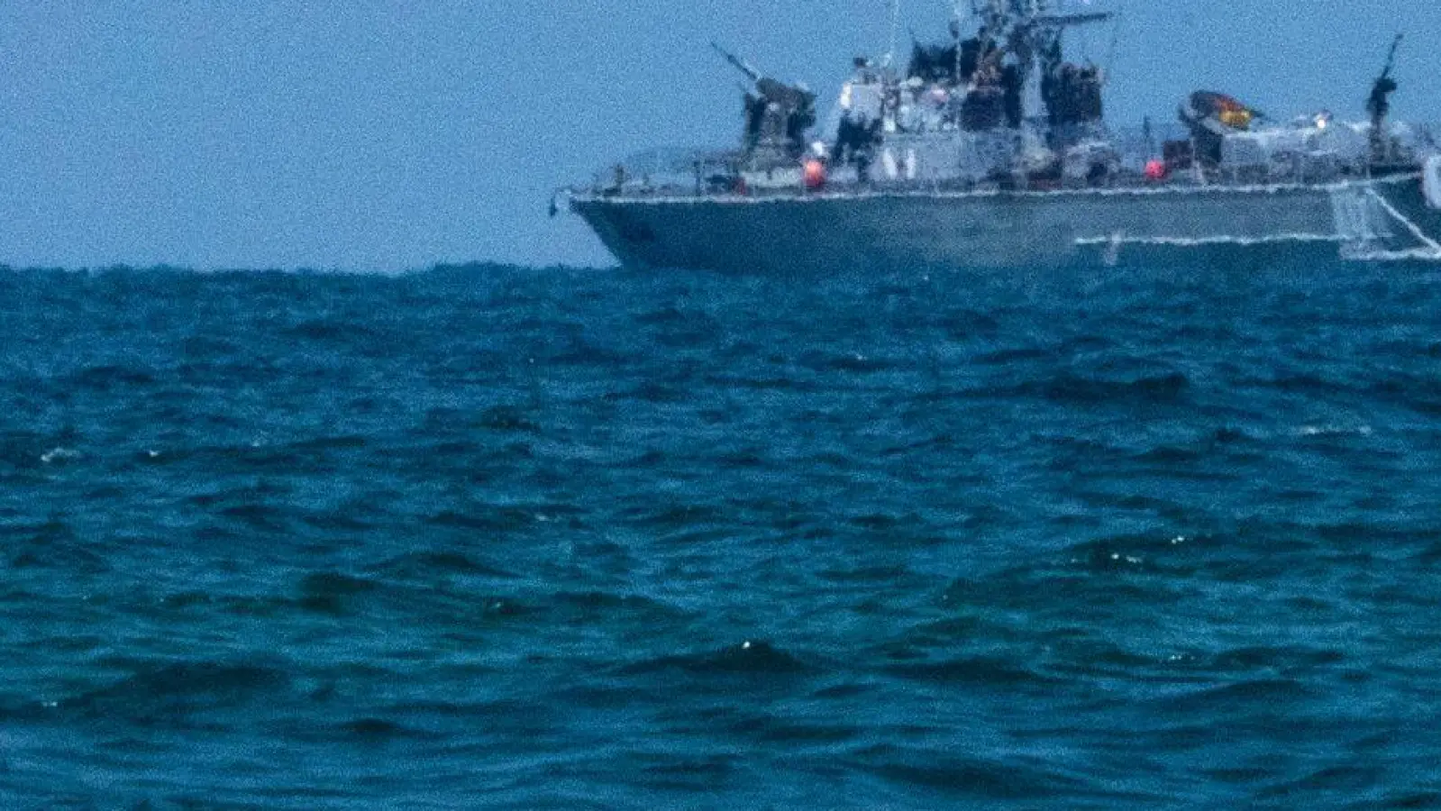 Ein Surfer wartet auf eine Welle, während ein israelisches Marineschiff im Mittelmeer vor der Küste von Hadera patrouilliert. (Foto: Ariel Schalit/AP)