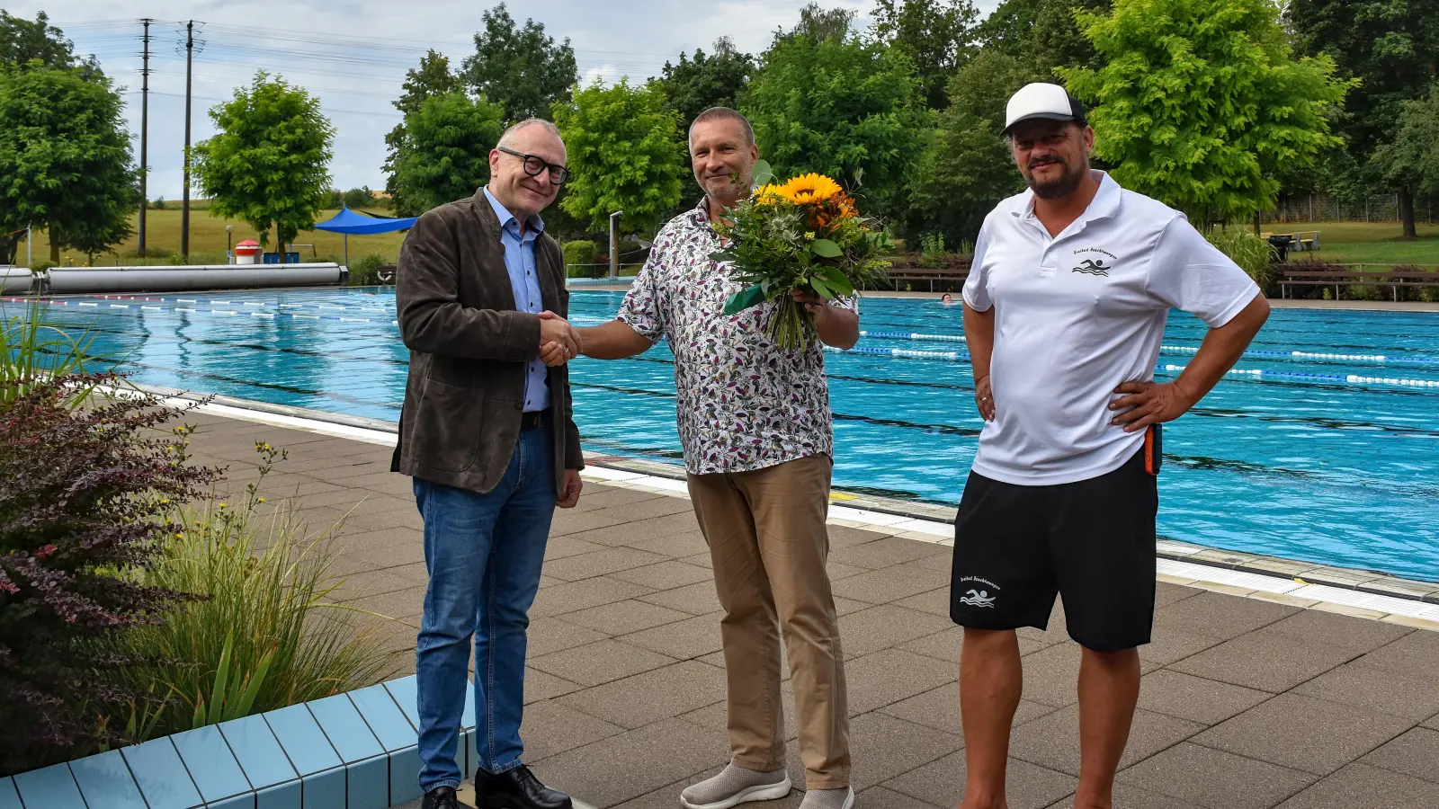 Bürgermeister Dr. Ulrich Grünwald (links) und Betriebsleiter Heiko Hercher (rechts) gratulierten Stammgast Wolfgang Kullmann mit einem Blumenstrauß und einem Gutschein für eine Saisonkarte. Marlies Kullmann war bei dem Termin verhindert. (Foto: Stadt Feuchtwangen/Rebecca Weber)
