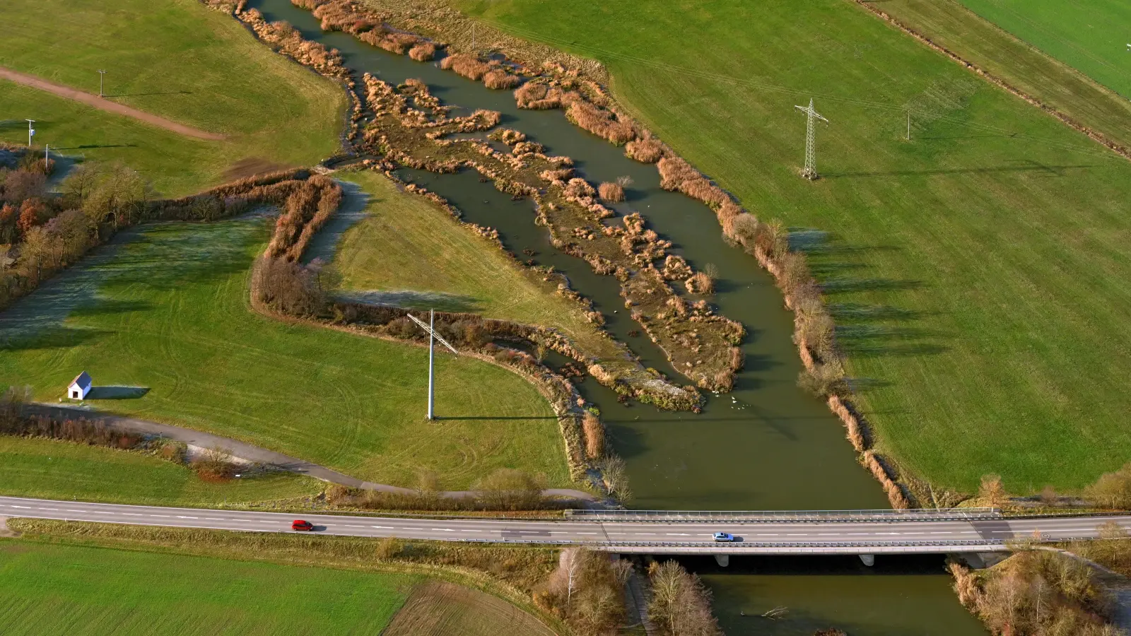 Das Foto zeigt die auf diesem von oben nach unten fließende renaturierte beziehungsweise umgestaltete Altmühl bei Ornbau mit der Staatsstraße 2411 Triesdorf-Arberg. Auf dem rechten Flussufer oben ist der Schilfgürtel zu sehen, der sich so stark ausgebreitet hat, dass etwa Fischern dort der Zugang zum Wasser stark erschwert wird. Das Schilf wird durch eine Beweidung mit Angus-Rindern eingedämmt. (Foto: Hajo Dietz)